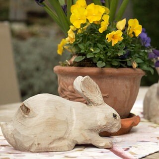 Zwei hölzerne Osterhasen sitzen auf einem Gartentisch. Zwischen ihnen ist ein Tontopf mit gelben und lianen Blumen.