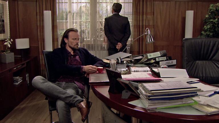 Constantin sitzt im Bürgermeisterbüro und trinkt Espresso, der Bürgermeister steht am Fenster und schaut raus