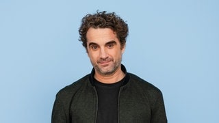 Der Schauspieler Oliver Wnuk trägt ein schwarzes Shirt und eine schwarze Jacke vor blauem Hintergrund und schaut direkt in die Kamera.