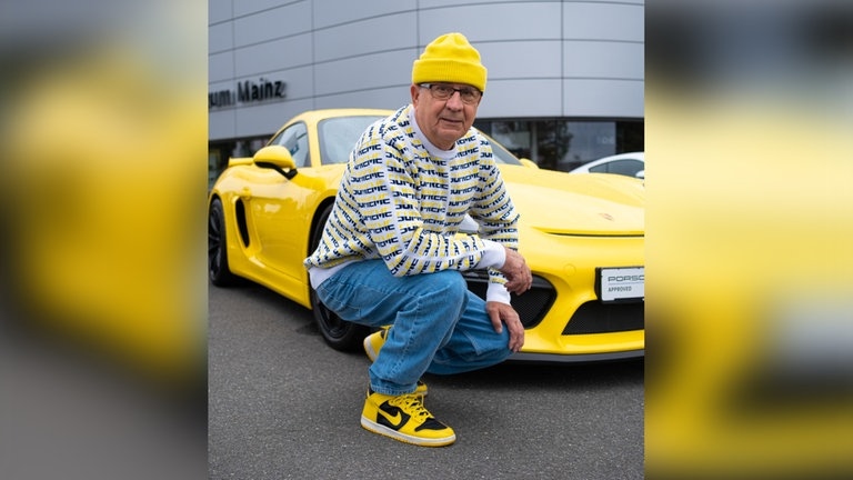 Ältere Person, die in Street-Style Klamotten vor einem Sportwagen posiert.