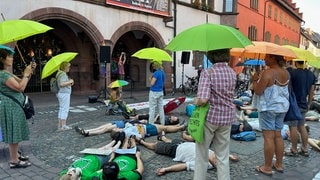 Menschen mit einem Regenschirm stehen vor dem Freiburger Rathaus. Andere liegen davor. Sie demonstrieren bei einer Kundgebung von Fridays for Future, was extreme Hitze mit Menschen macht.