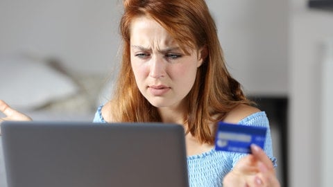 Frau sitzt mit Bankkarte in der Hand irritiert vor dem Laptop
