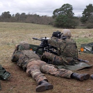 Mehrere Soldaten in Tarnanzügen sitzen auf einer Wiese. Einer von ihnen sitzt an einer großen Haubitze, ein anderer liegt links neben ihm. Rechts von ihnen sitzt ein Soldat mit einem Fernglas auf einem Stuhl.