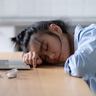 Symbolbild: Müde Studentin liegt vor ihrem Laptop auf dem Tisch und schläft