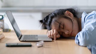 Symbolbild: Müde Studentin liegt vor ihrem Laptop auf dem Tisch und schläft