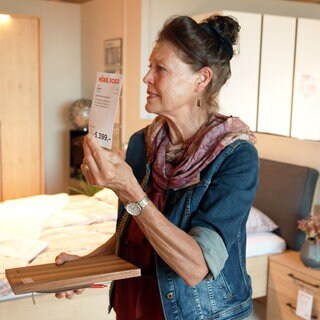 Verkäuferin Annette Schramm steht in einer Schlafzimmer-Nachbildung im Möbelhaus und hät ein Preisschild in die Luft. In der anderen Hand hält sie ein Klemmbrett.