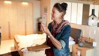Verkäuferin Annette Schramm steht in einer Schlafzimmer-Nachbildung im Möbelhaus und hät ein Preisschild in die Luft. In der anderen Hand hält sie ein Klemmbrett.