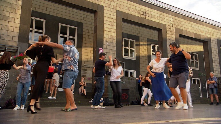 Mehrere Menschen tanzen gemeinsam auf einem Platz vor einem Haus.