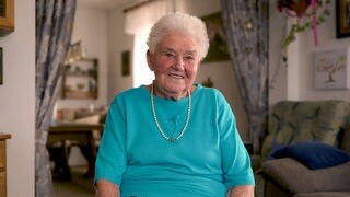 Rösle sitzt in ihrem Wohnzimmer. Sie trägt ein türkisfarbenes T-Shirt und eine Perlenkette. Sie hat kurze, graue Haare und lacht in die Kamera.