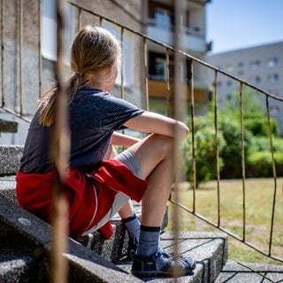 An die 3000 Kinder in Kaiserslautern leben in Bedarfsgemeinschaften, leiden unter Armut. (Symbolbild, Mädchen von hinten fotografiert, sitzt auf einer alten Treppe in einem Plattennbau-Wohngebiet)