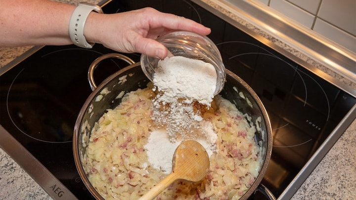 Gemäß Rezept wird Mehl in den Topf mit der Masse für den Zwiebelkuchen gegeben. Die Masse kommt dann auf den ausgerollten Hefeteig.