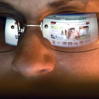 In der Brille einer jungen Frau spiegelt sich eine Internetseite zur Partnersuche.
