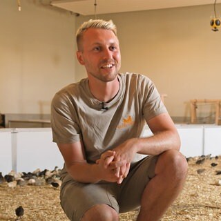 Michael Volk sitzt einem Stall. Im Hintergrund sieht man unscharf mehrere Wachteln auf dem Boden.