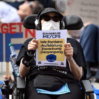 Demonstrantin und MECFS-Betroffene hält 2024 im Rollstuhl sitzend in Berlin ein Schild: "Wir fordern Aufklärung, Forschung, Versorgung". 1,5 Mio. Menschen in Deutschland haben Long Covid oder MECFS. Das erzeugt auch hohe Kosten für die Gesellschaft.