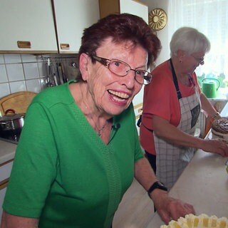 Trude und Lissy stehen in Lissys Küche und verzieren Kuchen. Trude ist im Vordergrund und lacht in die Kamera, Lissy steht hinter ihr an der Arbeitsplatte, vor ihr steht eine noch nicht verzierte Torte.