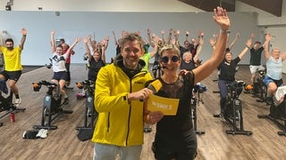 Silke und Benedikt vor dem Spinningkurs, der die Arme nach oben werfen vor Freude