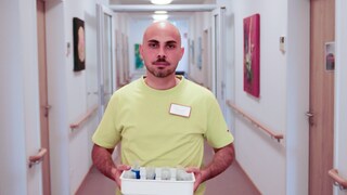 Alessandro geht mit einem Medikamenten-Tablett durch einen Flur in dem Altenheim, in dem er Pflegedienstleiter ist und schaut in die Kamera. Er ist Mitte 30, hat eine sportliche Statur, einen Drei-Tage-Bart, seine Arbeitskleidung sind eine weiße Hose zu einem grün-gelben Shirt.