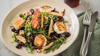 Ziegenkäsetaler auf Salat mit etwas Speck und Trauben, daneben Besteck und 