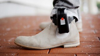 Eine elektronische Fußfessel ist oberhalb eines Schuhs an einem Bein befestigt.