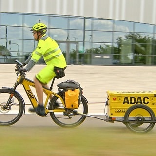 Heiko Graftl sitzt auf einem gelben Fahrrad mit einem Anhänger. Auf dem Anhänger steht ADAC. Er trägt eine Schutzweste und einen Helm und fährt nach links.