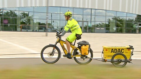 Heiko Graftl sitzt auf einem gelben Fahrrad mit einem Anhänger. Auf dem Anhänger steht ADAC. Er trägt eine Schutzweste und einen Helm und fährt nach links.