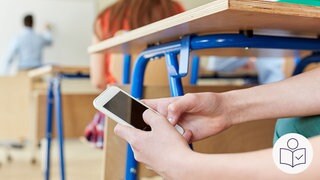 Ein Schüler hält ein Handy in der Hand. Der Schüler hält das Handy unter den Tisch. Der Schüler sitzt in einem Klassen·raum. In Rheinland-Pfalz sind Handys in der Schule jetzt verboten. Rechts das SWR Logo für Leichte Sprache.