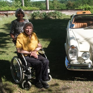 Oldtimersammler Rudolf Wagner sitzt im Rollstuhl. Hinter ihm steht seine Frau Lore. Die beiden stehen links von zwei von Rudolfs Oldtimern.