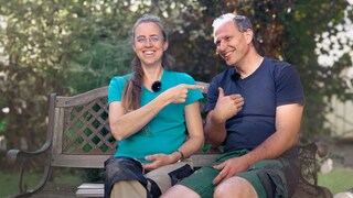  Das Paar Raphaela und Cornelius lacht herzlich miteinander auf einer Bank draußen im Freien. Die beiden haben zusammen sechs Söhne, eine richtige Großfamilie.
