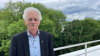 Hintergründig lächelnd lehnt Rammensee am Geländer eines Balkons, hinter ihm Bäume. Hans-Georg Rammensee forscht seit Jahren über das Immunsystem, und wie man es gegen Krebs einsetzen kann. 