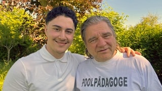 Mohamed und Eberhardt "Ebbe" Kögel stehen Arm in Arm auf einer Wiese im Stettener Freibad. Auf dem T-Shirt von "Ebbe" steht das Wort "Poolpädagoge". SWR1 Neuanfang. Mohamed: Flucht in ein neues Leben – ein junger Syrer schwimmt sich frei. Mohamed hat den syrischen Bürgerkrieg überlebt und fand eine neue Heimat in Stetten im Remstal. Warum ein Schwimmbad dabei entscheidend geholfen hat, erzählt er in SWR1 Neuanfang.