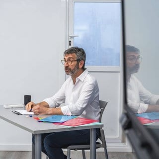 Mann mit Brille und Bart, der an einem modernen Schreibtisch mit bunten Ordnern sitzt