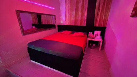 Zimmer in einer Prostituiertenwohnung in Lahnstein. Es ist spärlich beleuchtet, mit rotem Bett und Hygieneartikel auf dem Seitentisch. 