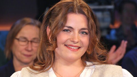 Ronja Forcher sitzt in einem Stuhl und hat eine weiße Bluse und eine beige Weste an