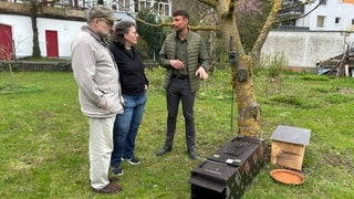 3 Personen stehen im Garten vor der Waschbären-Falle und unterhalten sich: Von links nach rechts Bernd Dauner, Monika Dauner und der Stadtjäger Julian Moosbauer.