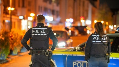 Eine Polizistin und ein Polizist stehen vor ihrem Polizeiwagen mit dem Rücken zur Kamera in einer Wohngegend. 