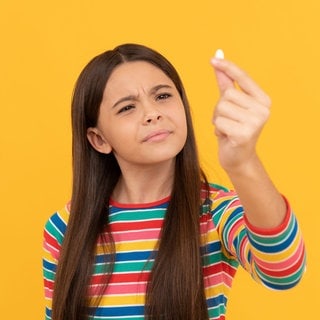 Mädchen hält skeptisch ein Dragee in die Höhe: Kinder brauchen für ihre gesunde Entwicklung Vitamine und Mineralstoffe. Aber was, wenn sie sie nicht über die Nahrung aufnehmen können oder wollen? Dann gibt es Nahrungsergänzungsmittel. Aber bei welchen Kindern ist die Nahrungsergänzung wirklich sinnvoll und welche Eltern sollten lieber die Finger davon lassen?