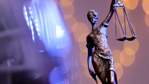Symbolbild. Eine Bronzestatue der Justitia steht auf einem Tisch. Im Hintergrund symbolisiertes Blaulicht. Ein 17-Jähriger sitzt wegen angeblicher sexueller Belästigung in Untersuchungshaft.