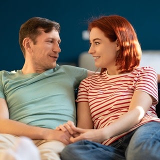 Ein junges Paar sitzt auf dem Sofa und lächelt sich an | Love Languages: So können sie euch helfen, eure Beziehung besser zu verstehen