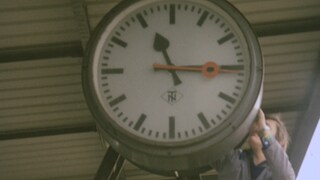 Eine große Uhr wird von einem Mann umgestellt. Er muss dafür auf eine Leiter steigen. 