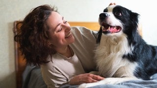 Eine Frau liegt mit ihrem Hund im Bett und lächelt ihn an. Eine neue Studie aus Wien hat untersucht, was im Hundehirn passiert, wenn Menschen lächeln.