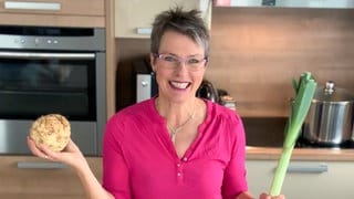 Karin ist Anti-Diät-Ernährungstherapeutin. Sie steht in ihrer Küche und hat einen Sellerie und einen Lauch in der Hand. Sie trägt ein pinkfarbenes Oberteil und lacht in die Kamera.