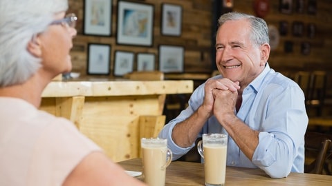 Mann und Frau sitzen sich im Café gegenüber und trinken Kaffee, er lächlt sie an. 