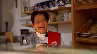Gerda steht in ihrem Dorfladen und lächelt, hinter hier liegen Brötchen und Brot im Regal. 