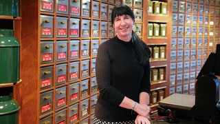 Margit steht hinter der Kasse in ihrem Tee-Fachgeschäft. Im Hintergrund stehen große Ladenregale gefüllt mit viel Tee. Sie lacht in die Kamera.