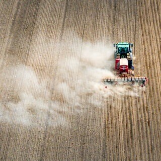 Ein Traktor zieht beim Ausbringen von Maissaat eine Drillmaschine über einen Acker und zieht eine große Staubwolke hinter sich her