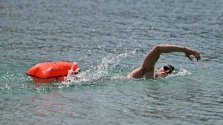 Ein Mann krault in einem See. Er zieht eine Schwimm-Boje hinter sich her. 