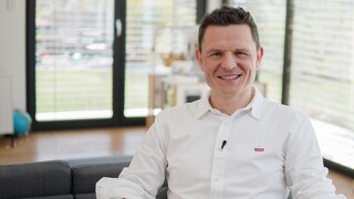 Achim sitzt in einem Stuhl. Er trägt ein weißes Hemd, hat eine sportliche Figur, ist nicht mehr untergewichtig und lächelt in die Kamera.