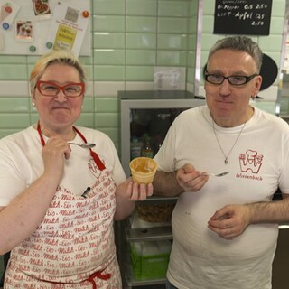 Jutta Wissenbach und ihr Mann Andreas Kalt stehen in ihrem Eiscafé. Jutta hält einen Eisbecher. Sie und ihr Mann haben jeweils einen Löffel in der Hand. Sie lächeln.