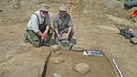 Zwei Männer sitzen auf einer archäologischen Grabung auf dem Boden. 