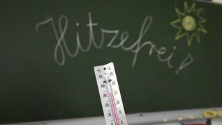 33 Grad werden auf einem Thermometer in einem Klassenzimmer einer Grundschule angezeigt. Auf einer Tafel im Hintergrund hat eine Lehrerin das Wort «Hitzefrei» geschrieben.